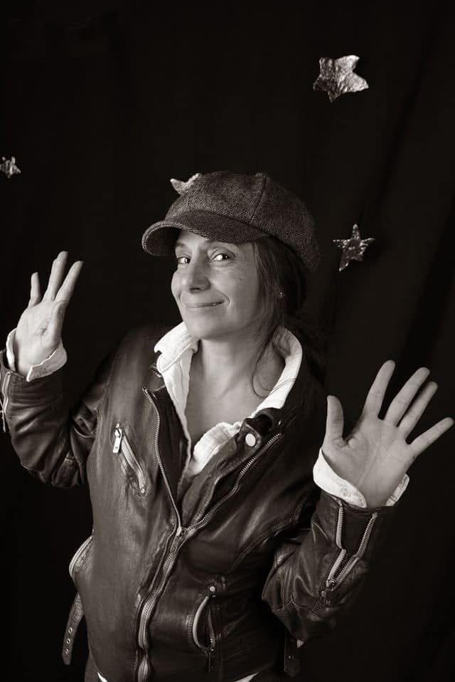 Portrait of the artist in a cap and leather jacket, hands raised, paper stars behind her